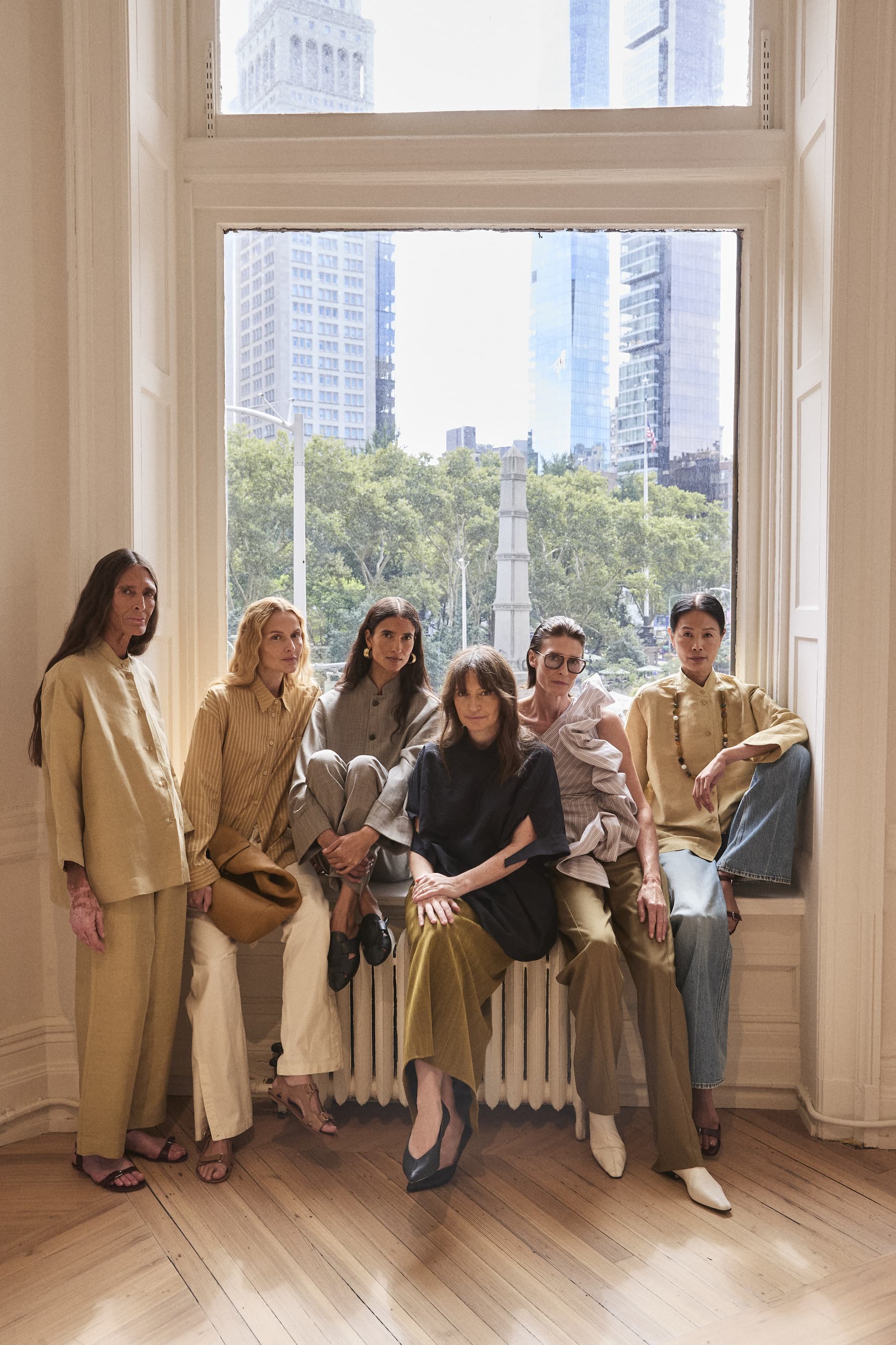 Five models posing in neutral-toned luxury apparel by a window with a New York City skyline view.