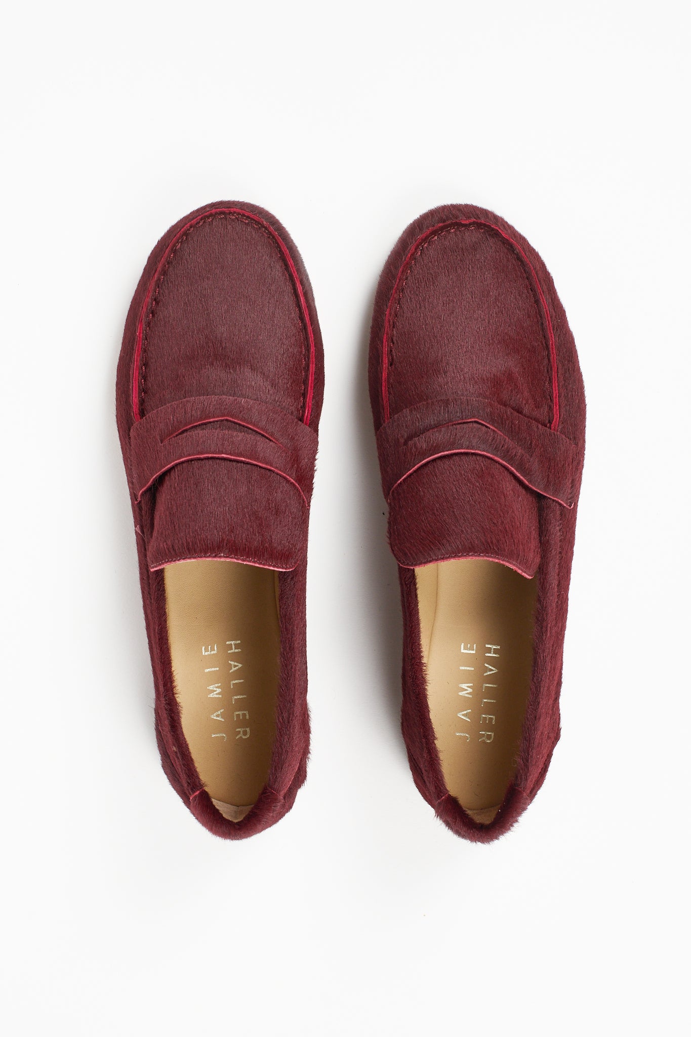 Top view of Jamie Haller claret burgundy ponyhair penny loafers with classic vamp straps, leather insoles, and rounded toes on a white background