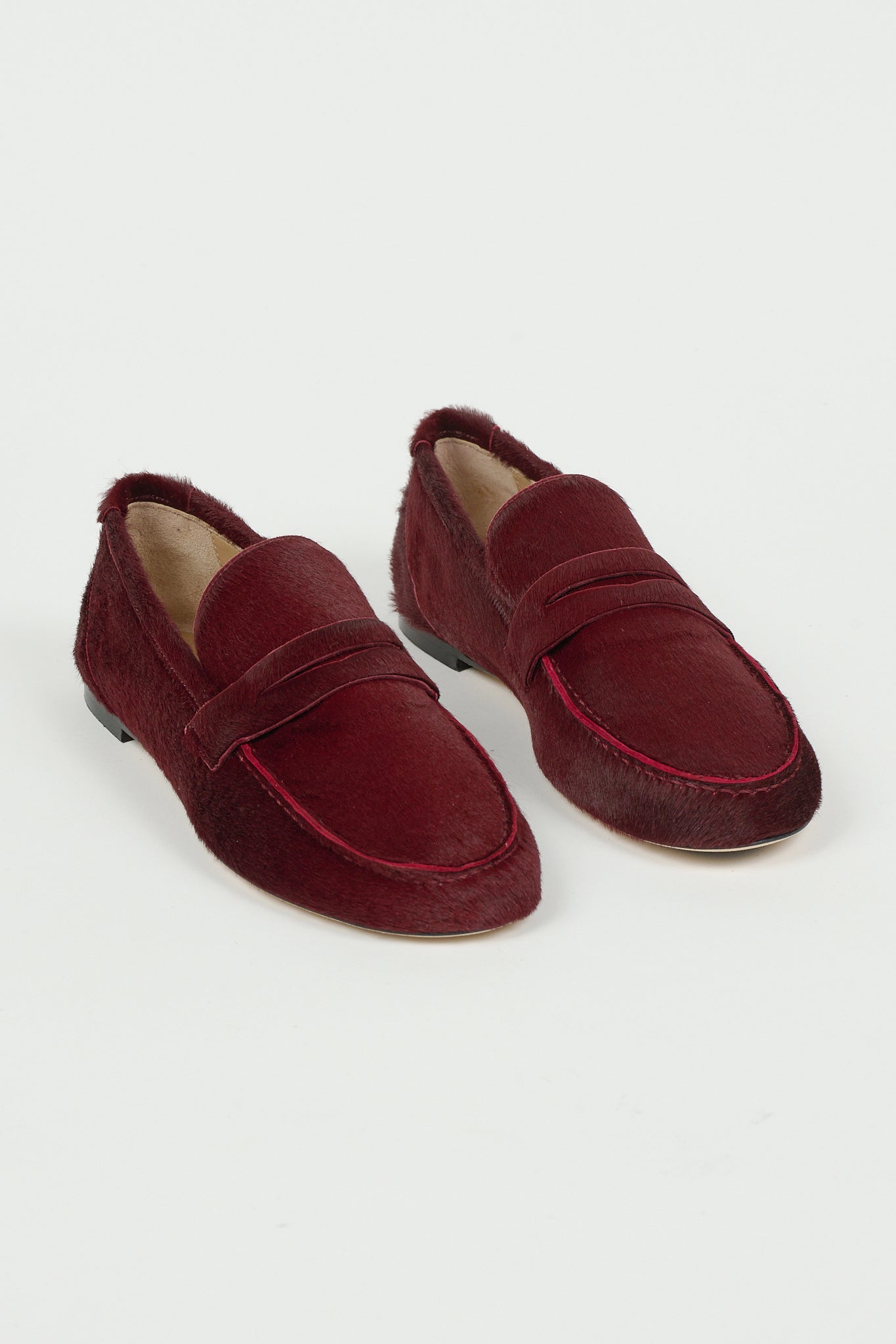 Pair of claret ponyhair penny loafers with moc toe stitching and leather soles, styled on a white background by Jamie Haller