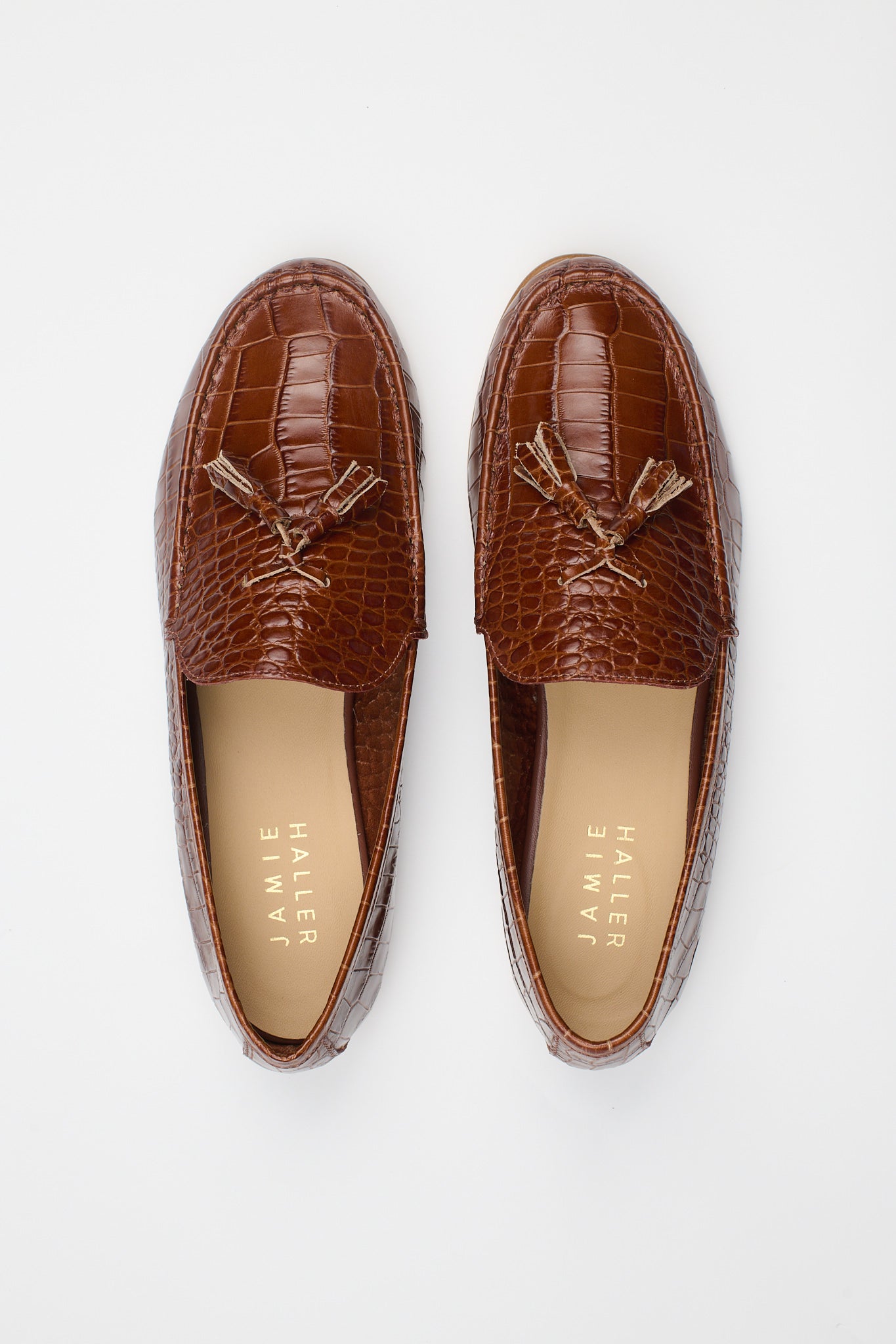 A pair of brown crocodile-embossed tassel loafers shown from a top-down view on a light gray background. The brand name 'JAMIE HALLER' is printed on the tan insoles.