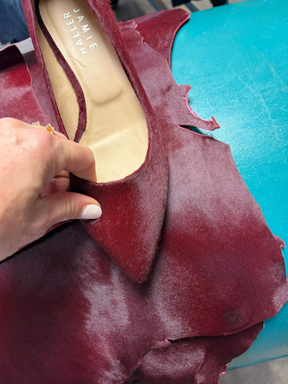 A close-up of a hand holding a burgundy suede shoe against a matching burgundy leather sheet, with a turquoise work surface in the background. The insole shows the brand name 'JAMIE HALLER'.