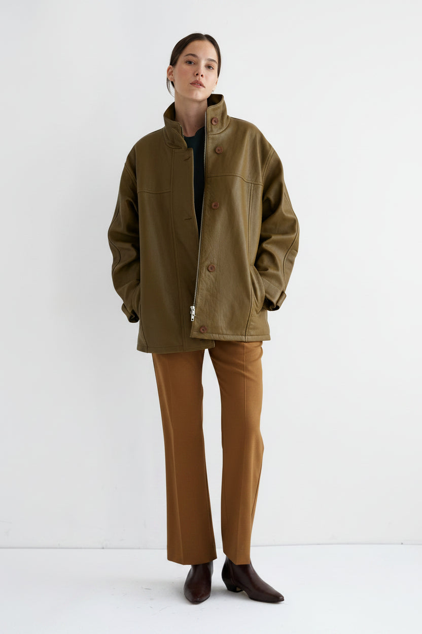 A model stands against a plain white studio background wearing an oversized olive green jacket over a dark top, tan pants, and dark brown shoes.