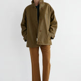 A model stands against a plain white studio background wearing an oversized olive green jacket over a dark top, tan pants, and dark brown shoes.