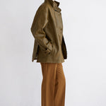 A model is shown in profile wearing an olive green jacket, camel brown wide-leg trousers, and dark brown leather boots against a clean white studio background.