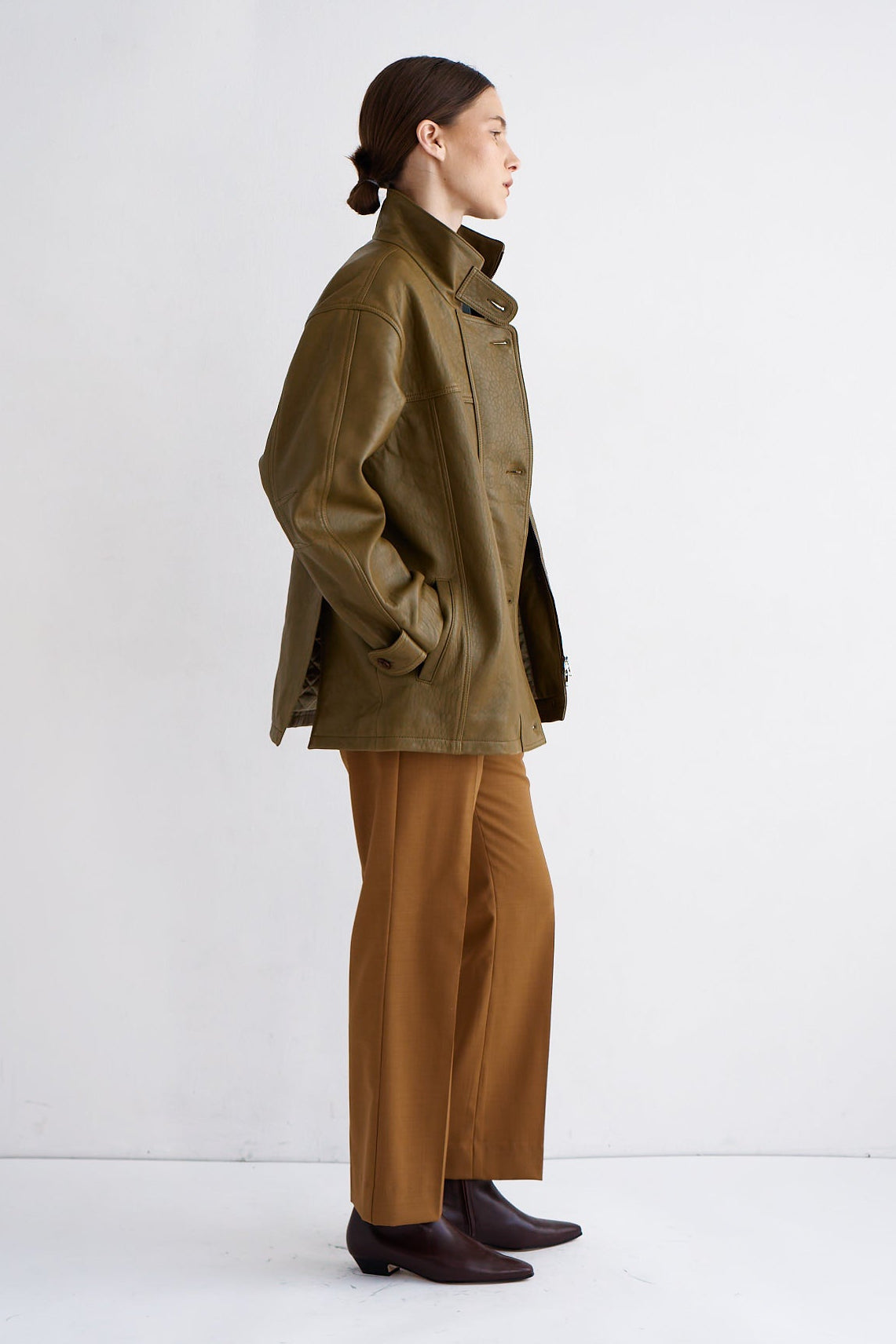 A model is shown in profile wearing an olive green jacket, camel brown wide-leg trousers, and dark brown leather boots against a clean white studio background.