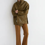 A model stands against a plain white studio wall wearing an oversized olive-brown jacket, brown trousers, and dark brown boots.