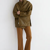 A model stands against a plain white studio wall wearing an oversized olive-brown jacket, brown trousers, and dark brown boots.