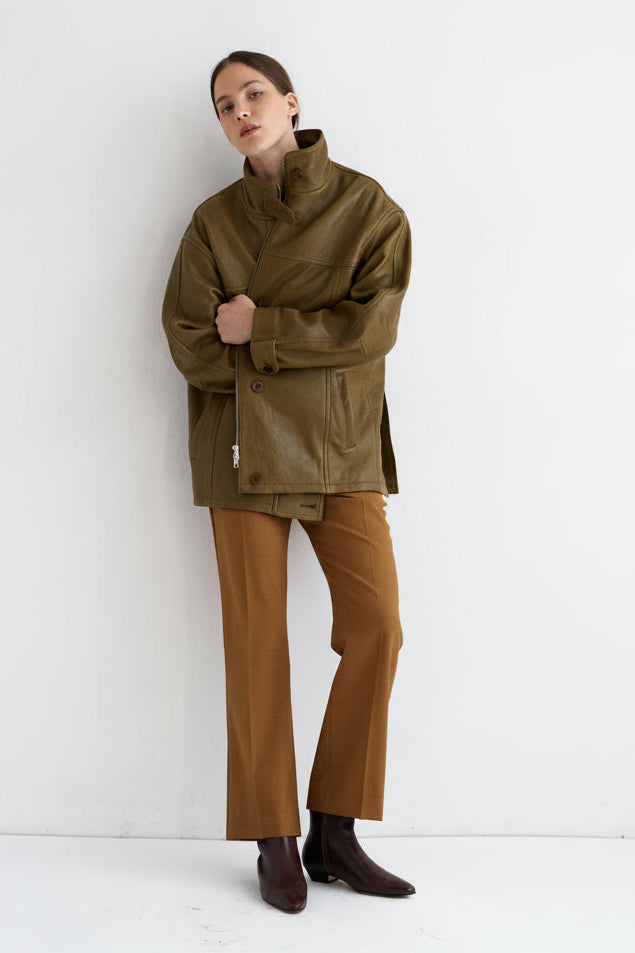 A model stands against a plain white studio wall wearing an oversized olive-brown jacket, brown trousers, and dark brown boots.
