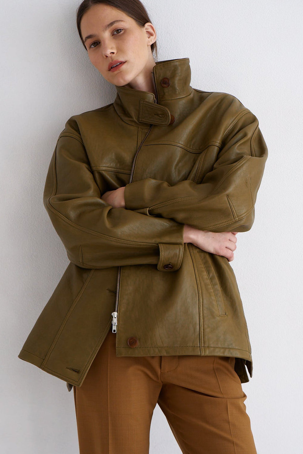 A model wearing an oversized olive green leather jacket with a high collar and brown trousers, posing against a plain light gray wall.