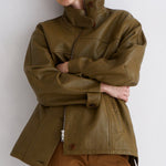 A model wearing an oversized olive green leather jacket with a high collar and brown trousers, posing against a plain light gray wall.
