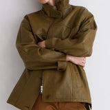 A model wearing an oversized olive green leather jacket with a high collar and brown trousers, posing against a plain light gray wall.