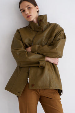 A model wearing an oversized olive green leather jacket with a high collar and brown trousers, posing against a plain light gray wall.