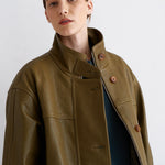 A woman wearing an olive green leather jacket with a high collar and brown buttons, layered over a dark blue top, photographed against a light gray studio background.