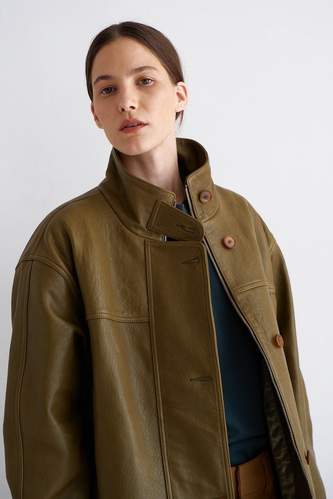 A woman wearing an olive green leather jacket with a high collar and brown buttons, layered over a dark blue top, photographed against a light gray studio background.