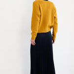 A woman is shown from behind wearing a mustard yellow knit sweater, a dark navy pleated midi skirt, and red flat shoes, set against a plain white studio background.