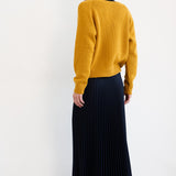 A woman is shown from behind wearing a mustard yellow knit sweater, a dark navy pleated midi skirt, and red flat shoes, set against a plain white studio background.