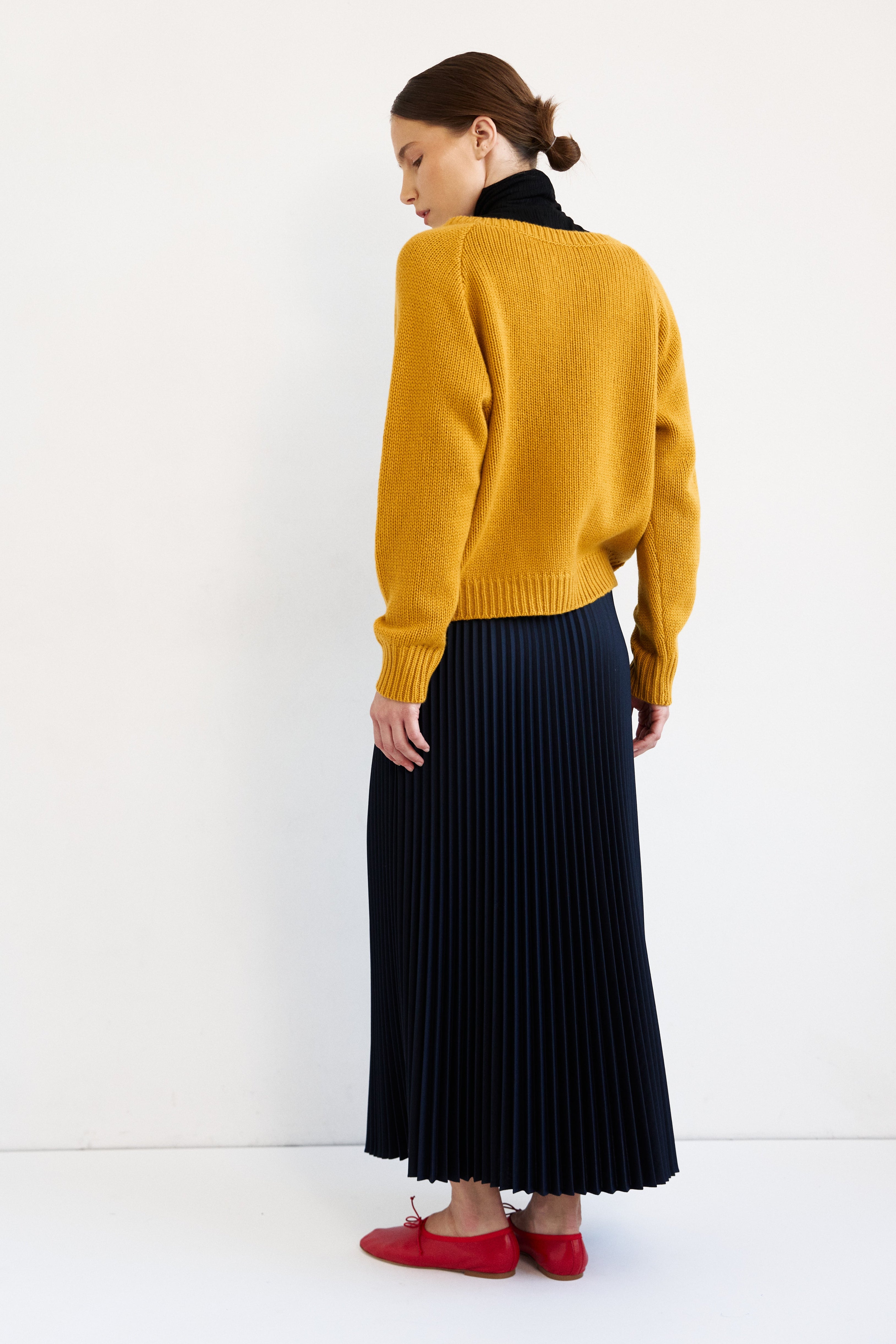 A woman is shown from behind wearing a mustard yellow knit sweater, a dark navy pleated midi skirt, and red flat shoes, set against a plain white studio background.