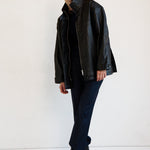A model stands in a studio wearing a black leather jacket over a black top and dark trousers, paired with black ankle boots, against a plain white background.