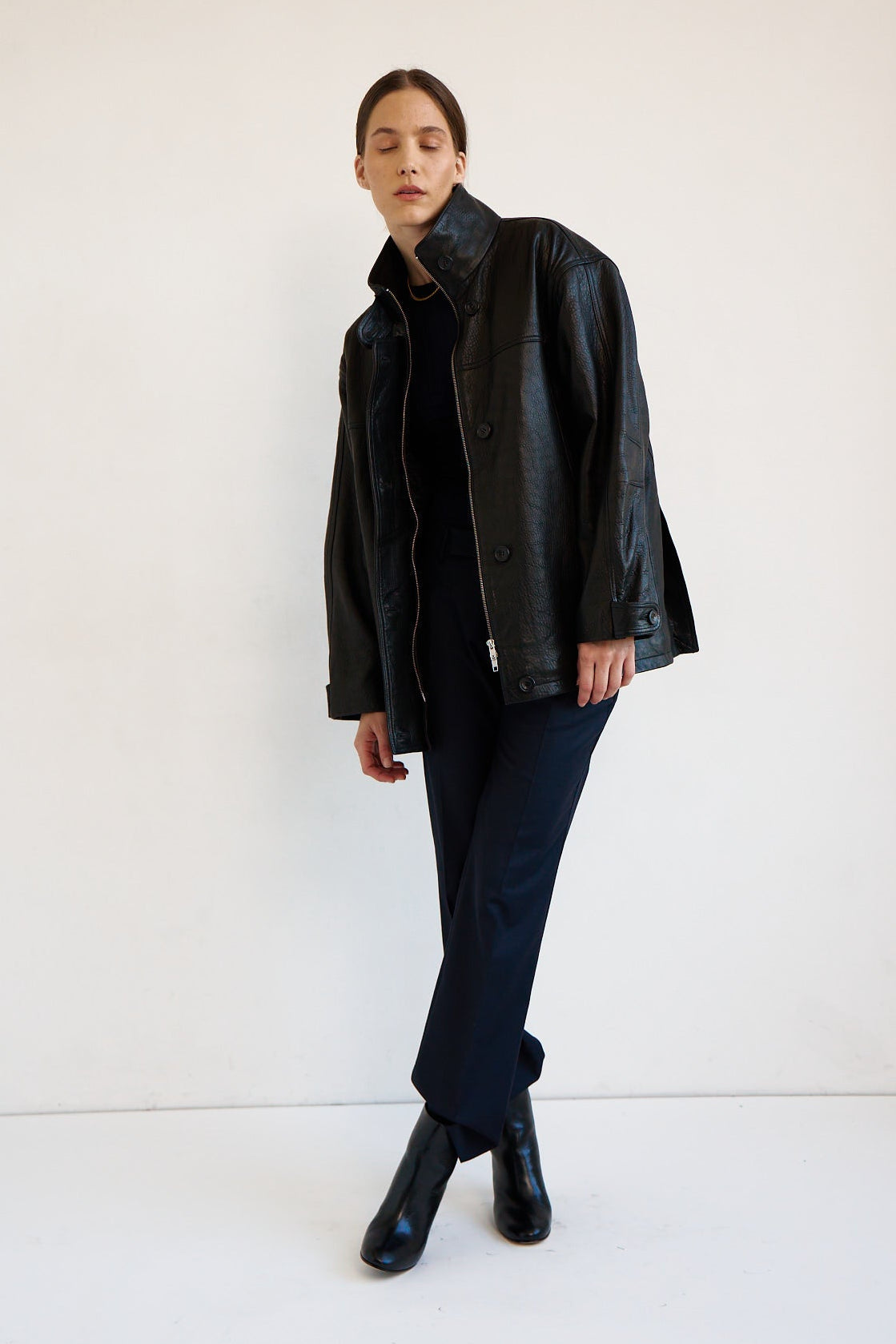 A model stands in a studio wearing a black leather jacket over a black top and dark trousers, paired with black ankle boots, against a plain white background.