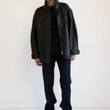 A model stands against a plain white studio background wearing a black leather jacket over a black top, dark navy pants, and black ankle boots.