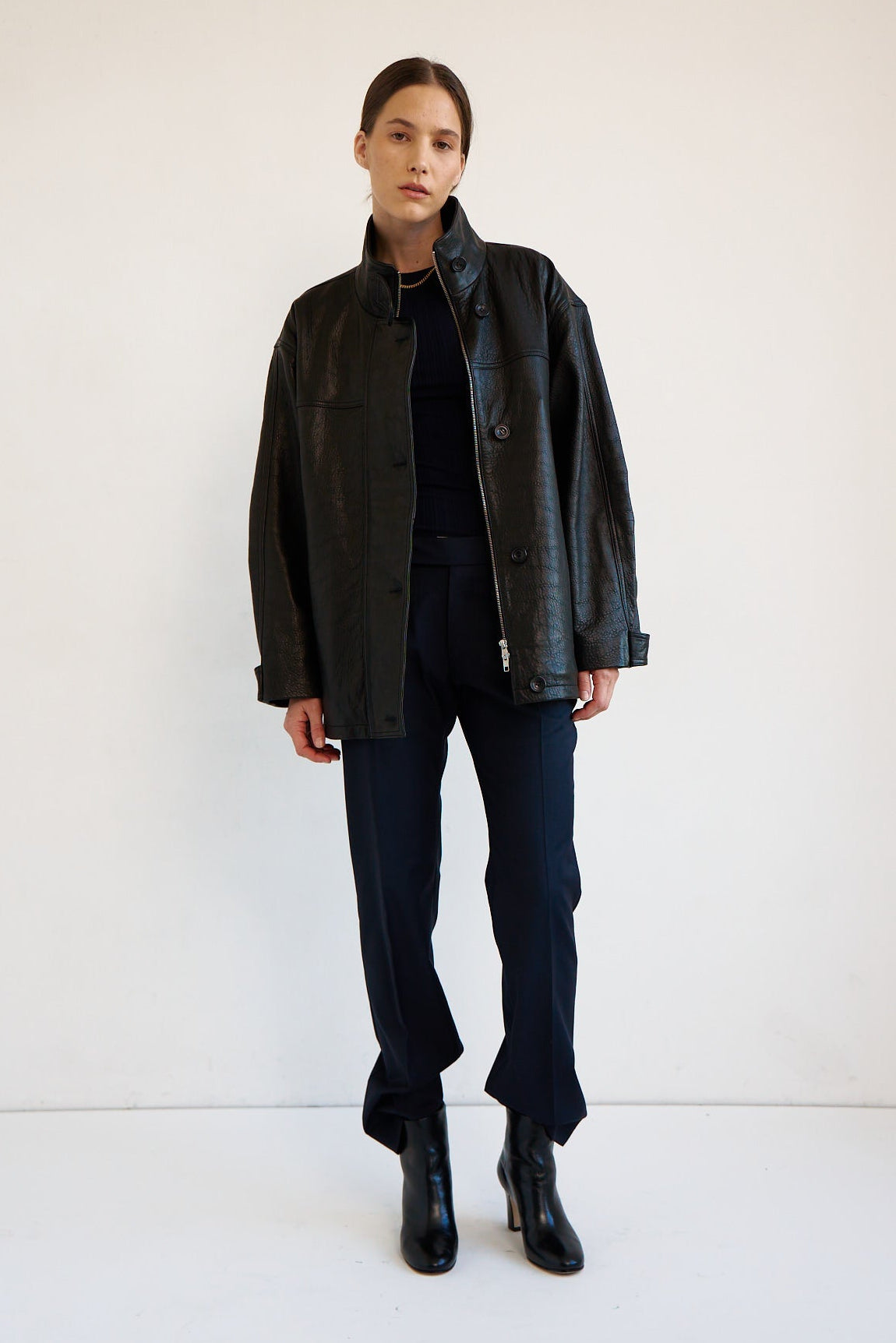 A model stands against a plain white studio background wearing a black leather jacket over a black top, dark navy pants, and black ankle boots.