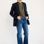A model stands against a plain white studio background wearing a dark navy blazer, a green and gold striped shirt, blue jeans, and brown shoes.