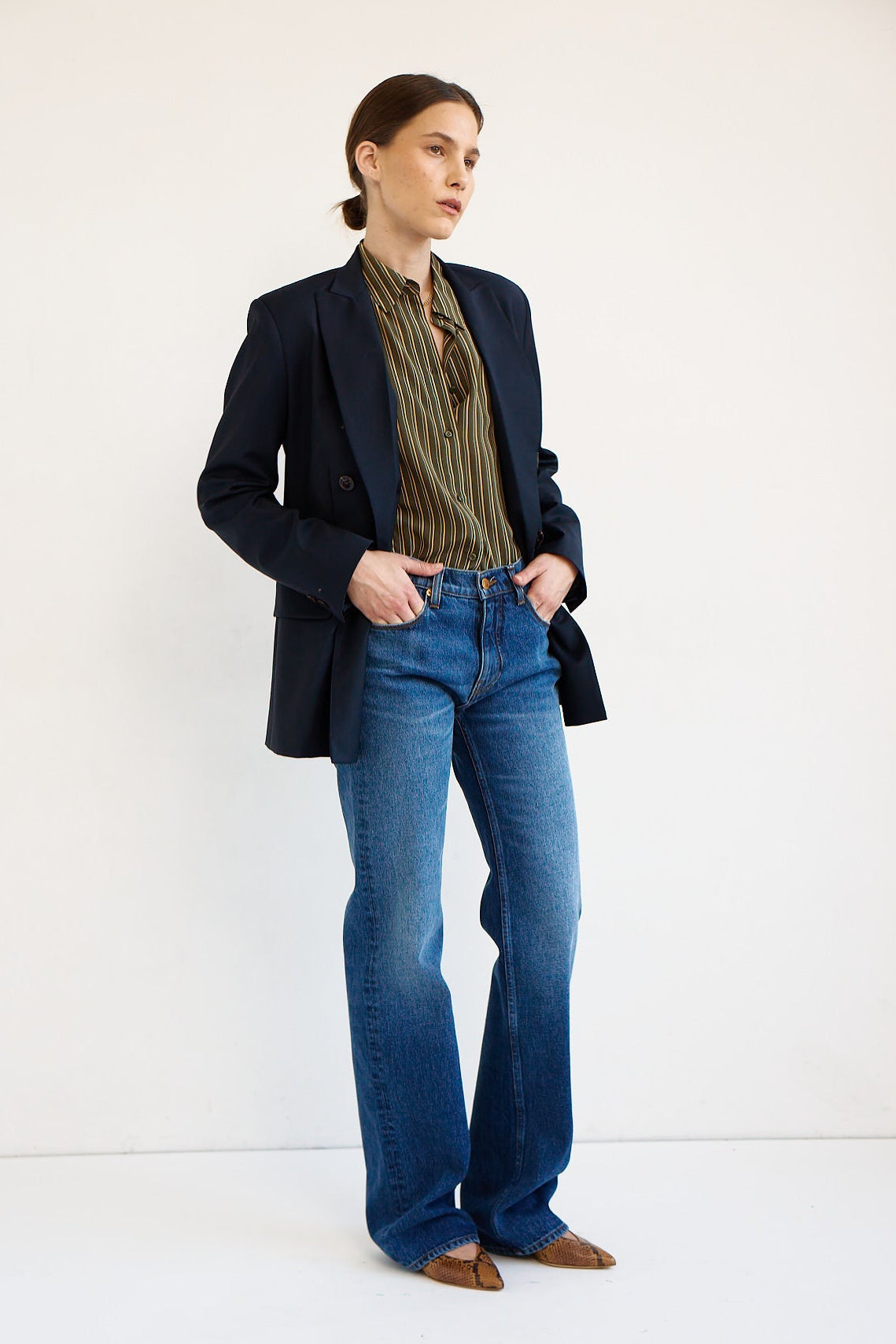 A model stands against a plain white studio background wearing a dark navy blazer, a green and gold striped shirt, blue jeans, and brown shoes.