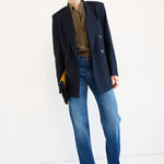 A model stands against a plain white studio background wearing an oversized dark navy blazer over a brown and gold striped shirt, blue jeans, and brown pointed shoes, while holding a small clutch.
