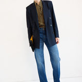 A model stands against a plain white studio background wearing an oversized dark navy blazer over a brown and gold striped shirt, blue jeans, and brown pointed shoes, while holding a small clutch.