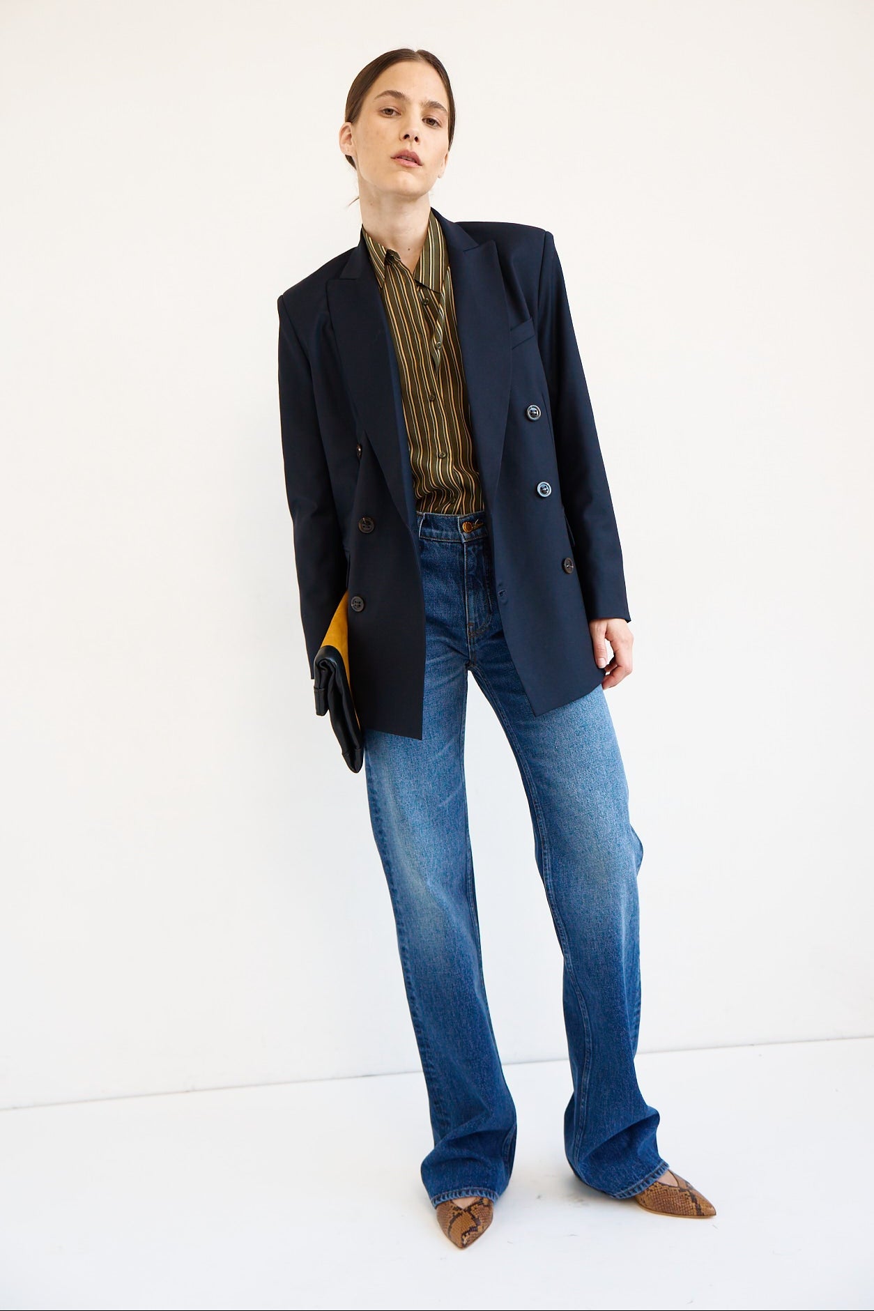 A model stands against a plain white studio background wearing an oversized dark navy blazer over a brown and gold striped shirt, blue jeans, and brown pointed shoes, while holding a small clutch.