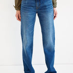 A person wearing blue straight-leg denim jeans with a subtle wash fade, paired with a striped long-sleeve shirt and brown pointed-toe shoes, shown from the waist down against a plain white studio background.