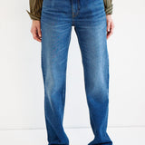 A person wearing blue straight-leg denim jeans with a subtle wash fade, paired with a striped long-sleeve shirt and brown pointed-toe shoes, shown from the waist down against a plain white studio background.