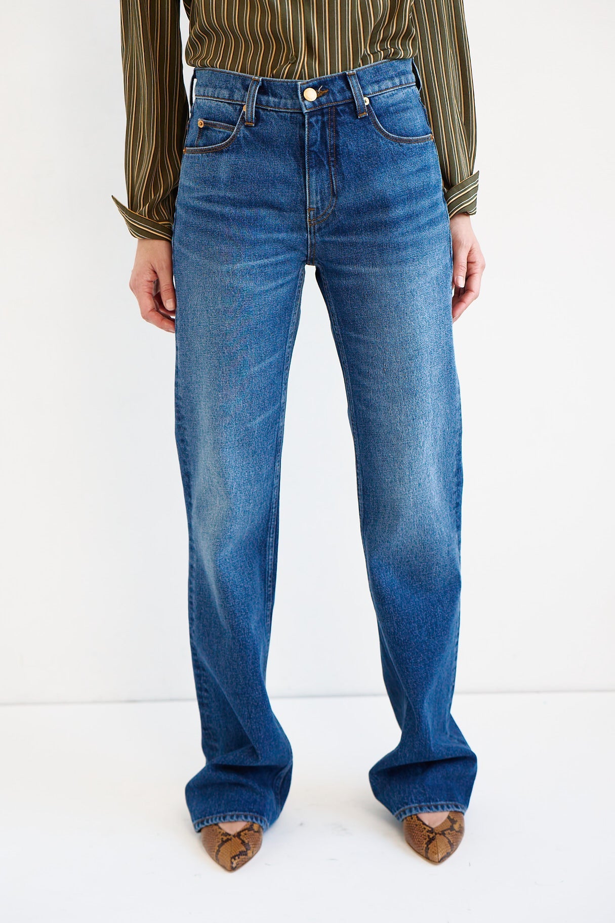 A person wearing blue straight-leg denim jeans with a subtle wash fade, paired with a striped long-sleeve shirt and brown pointed-toe shoes, shown from the waist down against a plain white studio background.