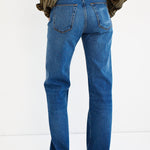 Rear view of a person wearing blue straight-leg jeans with a striped shirt and brown shoes, standing against a plain white studio background.