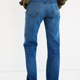 Rear view of a person wearing blue straight-leg jeans with a striped shirt and brown shoes, standing against a plain white studio background.