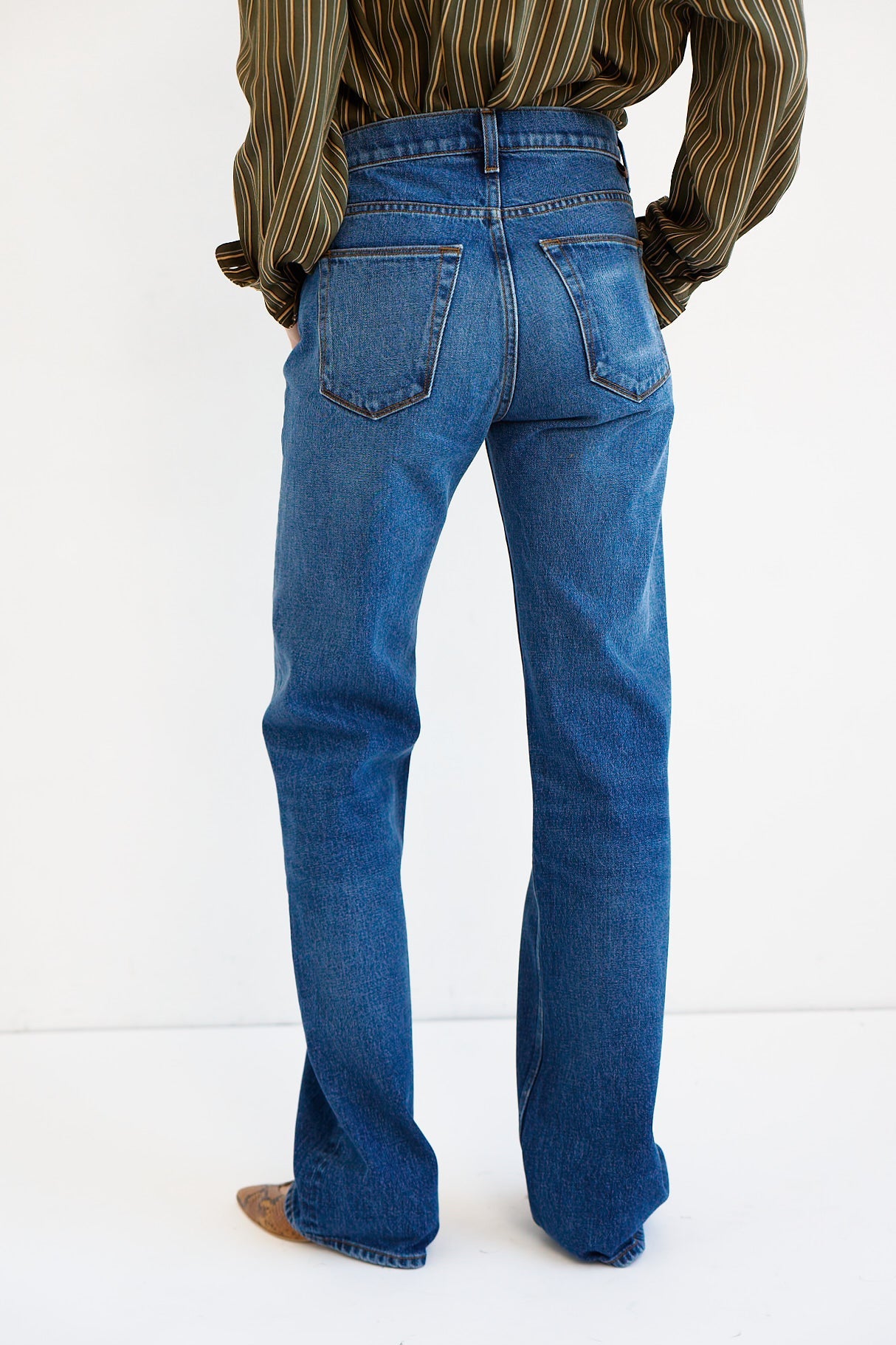 Rear view of a person wearing blue straight-leg jeans with a striped shirt and brown shoes, standing against a plain white studio background.
