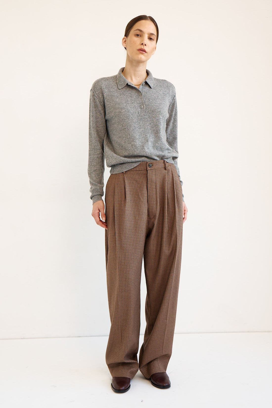 A model stands against a plain white studio background wearing a gray long-sleeve knit polo shirt and high-waisted brown wide-leg trousers.