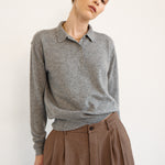 A woman wearing a heather gray long-sleeve polo shirt tucked into brown patterned trousers, photographed against a plain off-white studio background.