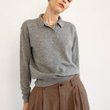 A woman wearing a heather gray long-sleeve polo shirt tucked into brown patterned trousers, photographed against a plain off-white studio background.