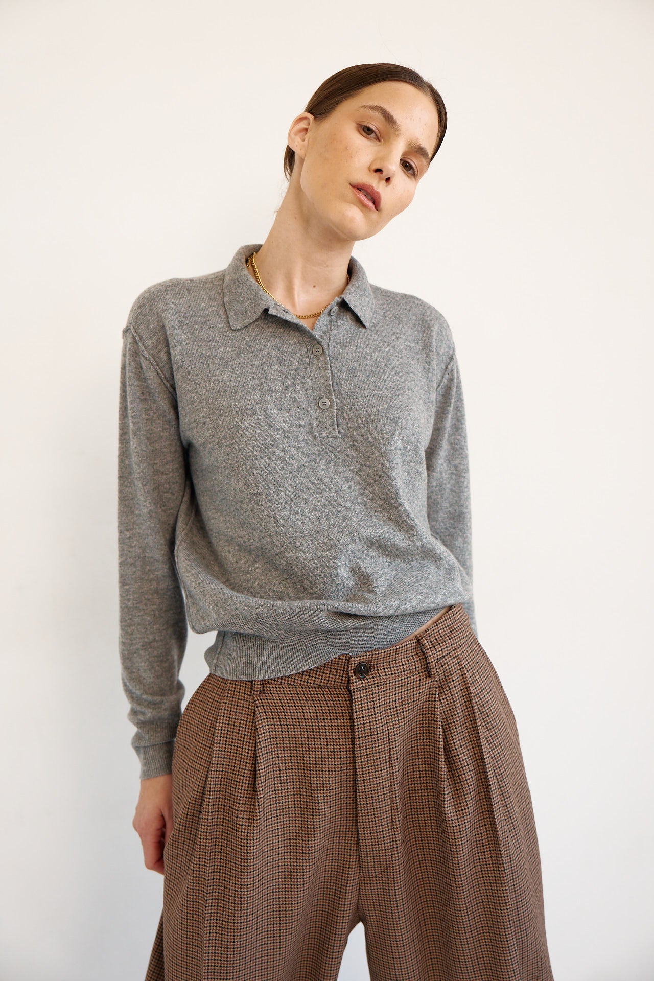 A woman wearing a heather gray long-sleeve polo shirt tucked into brown patterned trousers, photographed against a plain off-white studio background.