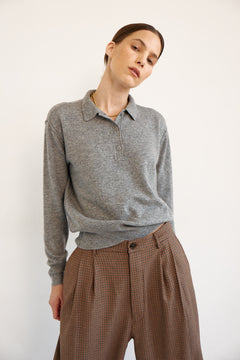 A woman wearing a heather gray long-sleeve polo shirt tucked into brown patterned trousers, photographed against a plain off-white studio background.