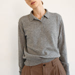 A woman wearing a heather gray long-sleeve polo shirt and brown plaid trousers, photographed against a plain off-white background.