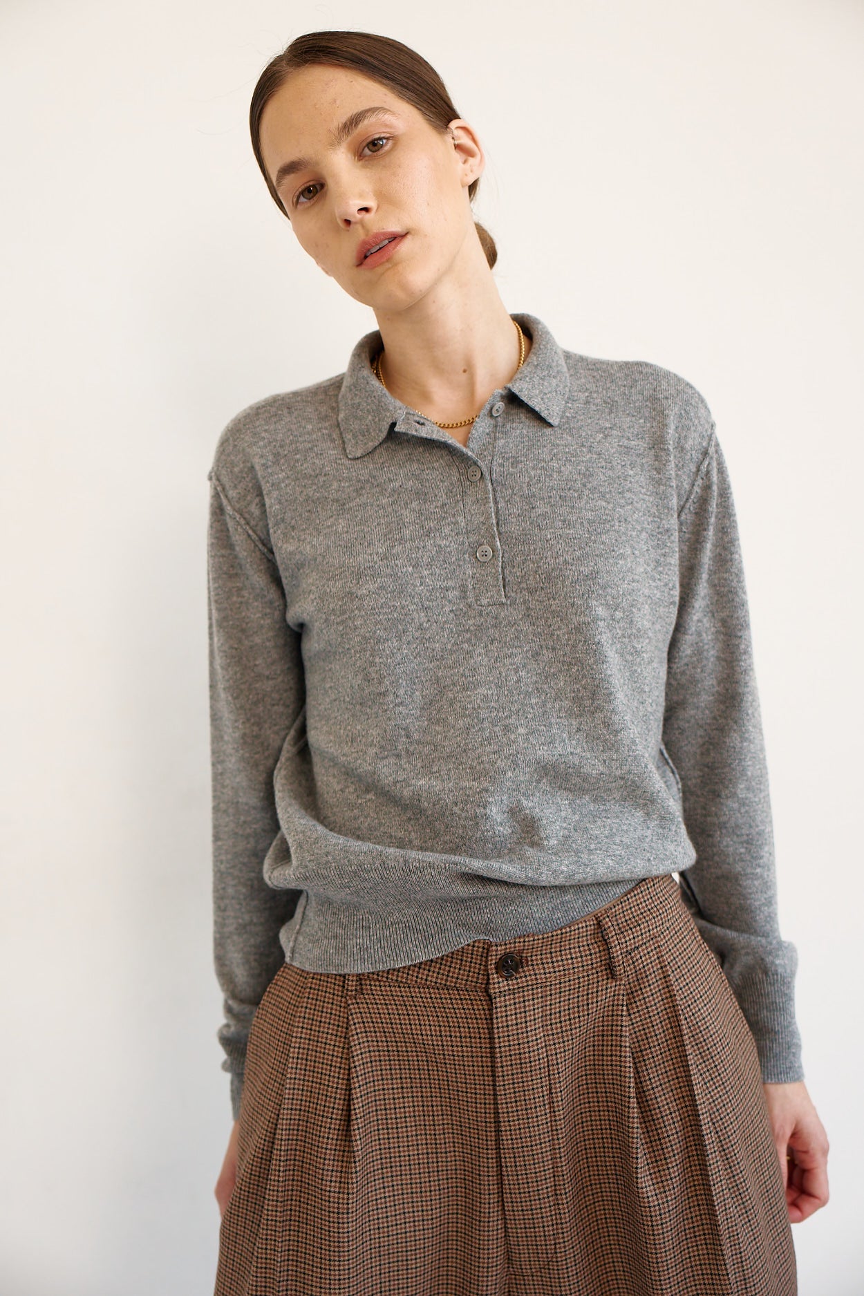 A woman wearing a heather gray long-sleeve polo shirt and brown plaid trousers, photographed against a plain off-white background.