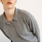 A close-up shot of a model wearing a heather gray long-sleeve polo shirt and brown plaid trousers, accessorized with a delicate gold chain necklace, against a plain off-white background.