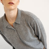 A close-up shot of a model wearing a heather gray long-sleeve polo shirt and brown plaid trousers, accessorized with a delicate gold chain necklace, against a plain off-white background.