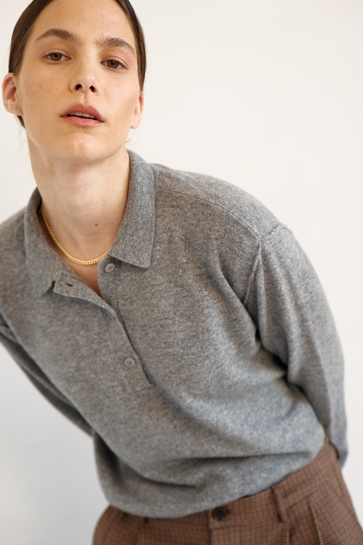 A close-up shot of a model wearing a heather gray long-sleeve polo shirt and brown plaid trousers, accessorized with a delicate gold chain necklace, against a plain off-white background.