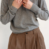A person wearing a gray long-sleeve knit polo shirt and brown checked pleated trousers, with hands adjusting the collar and button.