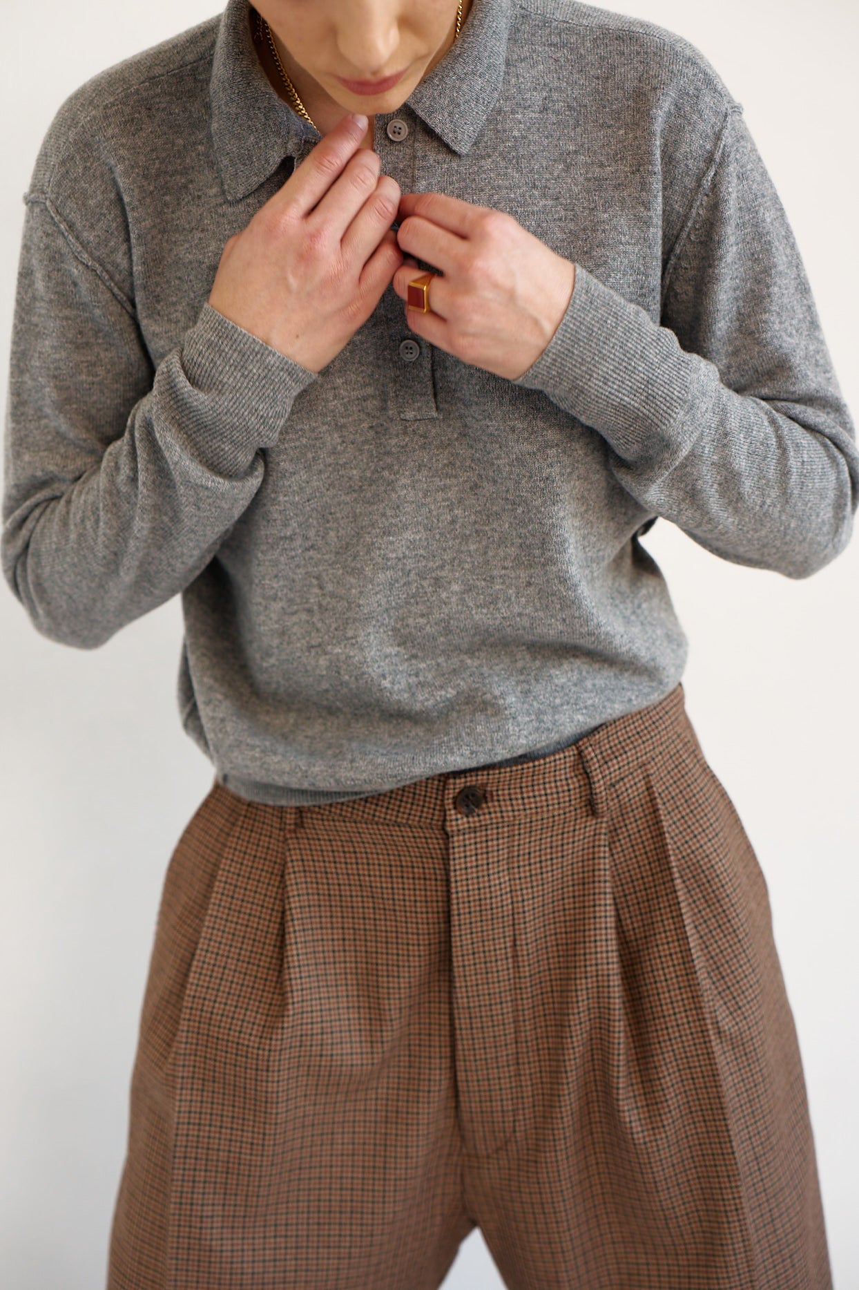 A person wearing a gray long-sleeve knit polo shirt and brown checked pleated trousers, with hands adjusting the collar and button.