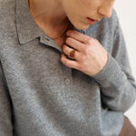 A close-up lifestyle shot of a model wearing a gray knit polo sweater, a gold chain necklace, and a gold ring with a red stone.