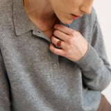 A close-up lifestyle shot of a model wearing a gray knit polo sweater, a gold chain necklace, and a gold ring with a red stone.